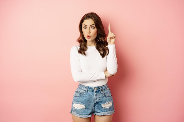Jonge brunette glam girl die vinger opsteekt, omhoog wijst, promo-aanbieding laat zien, eureka-gebaar, iets realiseert, staande over roze achtergrond