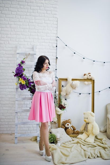 Jonge brunette meisje in roze rok en witte blouse poseerde binnen tegen kamer met speelgoed beer
