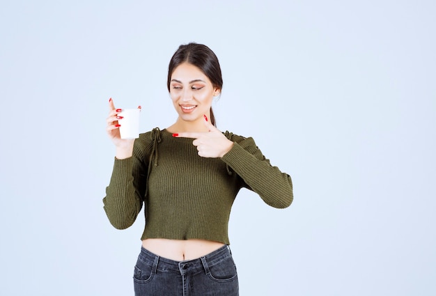 Jonge brunette vrouw die plastic beker gelukkig vasthoudt en ernaar wijst.