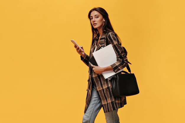 Jonge brunette vrouw in oversized jas en spijkerbroek houdt telefoon en witte notebooks Aantrekkelijk meisje met zwarte tas poseert op oranje achtergrond