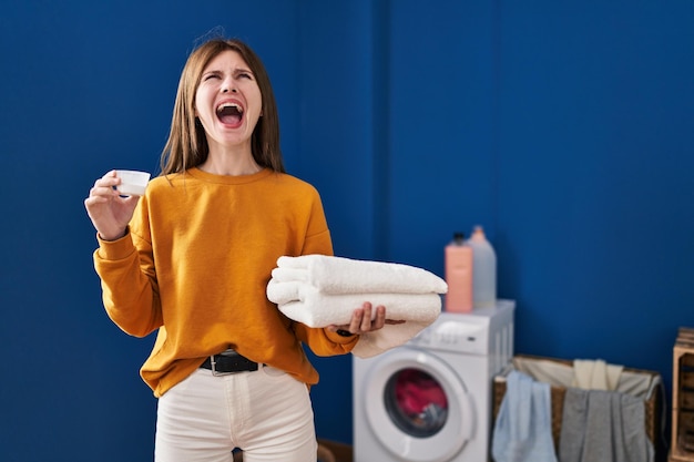 Gratis foto jonge brunette vrouw met wasmiddel en schone was boos en gek schreeuwend gefrustreerd en woedend schreeuwend van woede die omhoog kijkt