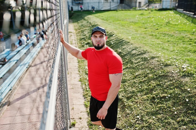 Jonge brute, bebaarde gespierde man draagt een rood shirt, korte broek en pet in het stadion