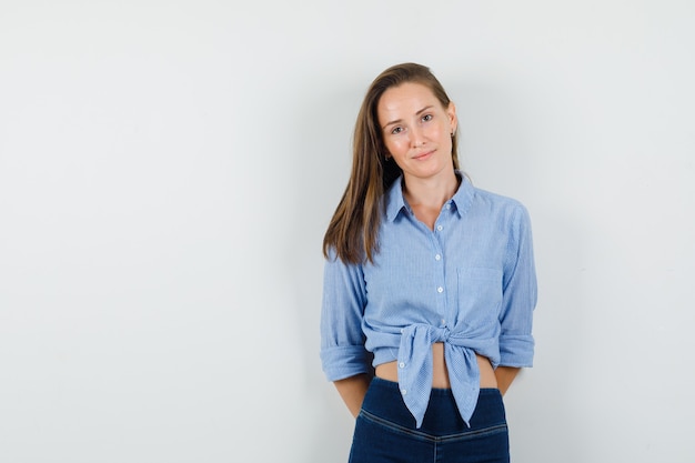 Jonge dame camera kijken met handen op haar rug in blauw shirt, broek en vrolijk kijken