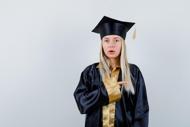 Jonge dame die camera in academische kleding richt en verbaasd kijkt.