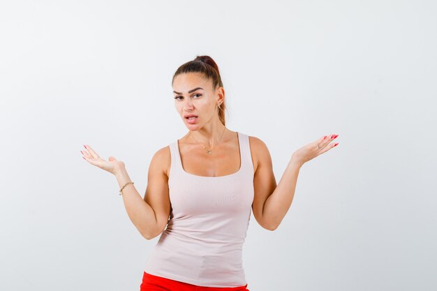 Jonge dame die de handpalmen opzij spreidt in beige tanktop en besluiteloos kijkt, vooraanzicht.