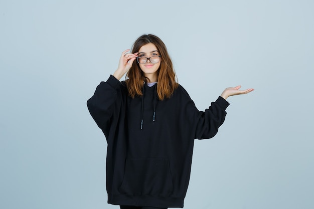 Gratis foto jonge dame die de vingers op een bril houdt terwijl ze doet alsof ze iets laat zien in een oversized hoodie, broek en er vrolijk uitziet, vooraanzicht.