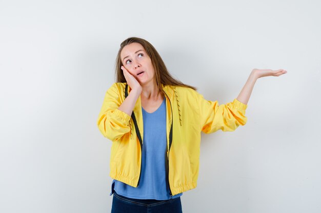 Jonge dame die doet alsof ze iets laat zien, wang op palm leunend in t-shirt, jasje en peinzend kijkend. vooraanzicht.