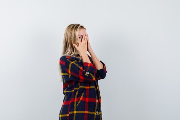 Jonge dame die door vingers kijkt terwijl handen op gezicht in geruit overhemd kijkt en schattig, vooraanzicht kijkt.