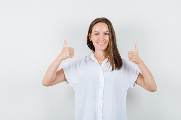 Jonge dame die duim toont terwijl het glimlachen in witte blouse en gelukkig kijkt.