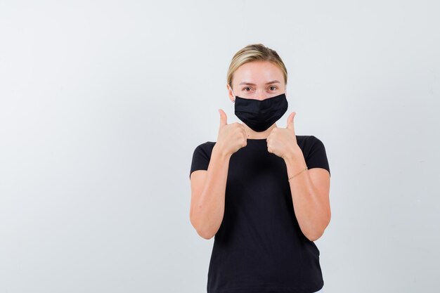 Jonge dame die duimen laat zien in t-shirt, broek, medisch masker en er vrolijk uitziet