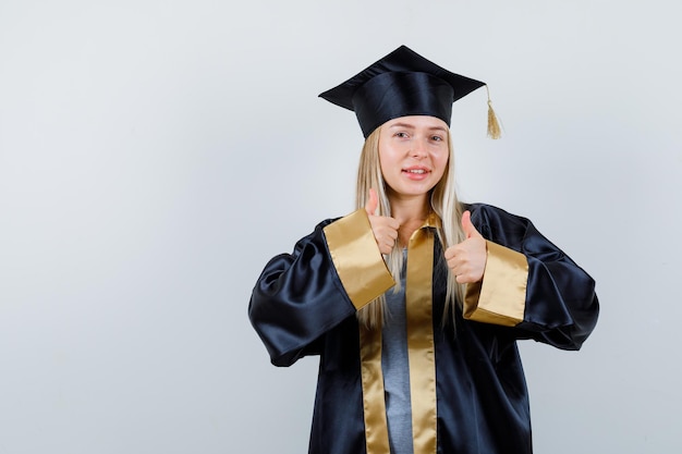 Gratis foto jonge dame die duimen opduikt in academische kleding en er zelfverzekerd uitziet.