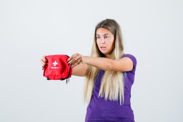 Jonge dame die EHBO-doos in violet t-shirt toont en verbaasd, vooraanzicht kijkt.