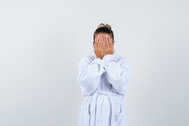 Jonge dame die haar gezicht bedekt met de handen in een badjas en er zelfverzekerd uitziet