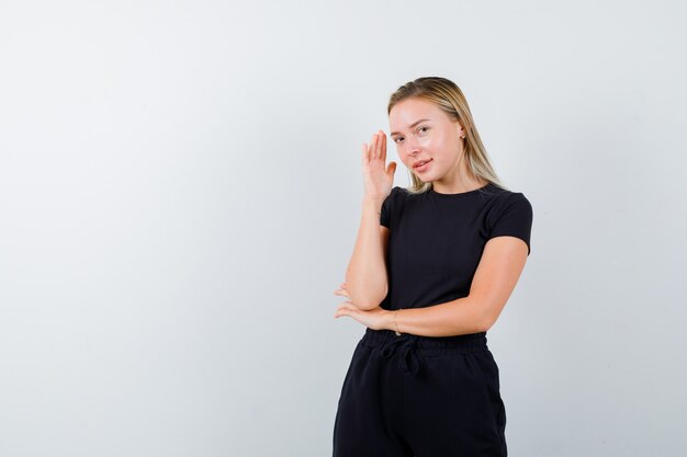 Jonge dame die hand dichtbij gezicht in t-shirt, broek houdt en vrolijk kijkt. vooraanzicht.