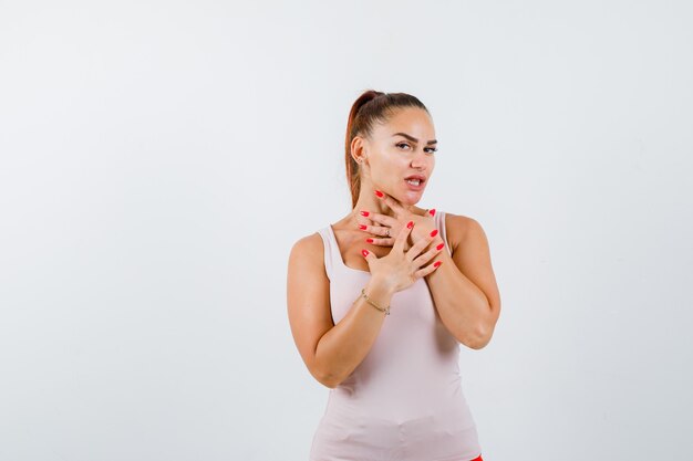 Jonge dame die hand op keel en borst in beige tanktop houdt en verward kijkt