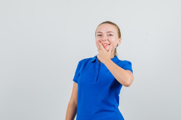 Jonge dame die hand op kin in blauw t-shirt houdt en gelukkig kijkt