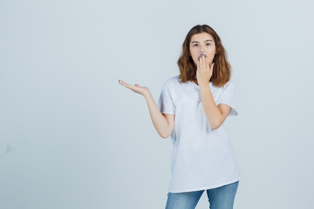 Jonge dame die hand op mond houdt terwijl iets in t-shirt, jeans wordt getoond en verbaasd, vooraanzicht kijkt.