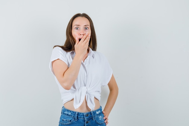 Jonge dame die hand op mond houdt terwijl verrassend in witte blouse, vooraanzicht.