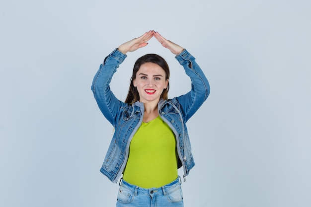 Jonge dame die het gebaar van het huisdak in denimoutfit toont en er vrolijk uitziet, vooraanzicht.