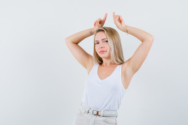 Jonge dame die hoorngebaar met vingers in witte blouse toont en gek kijkt