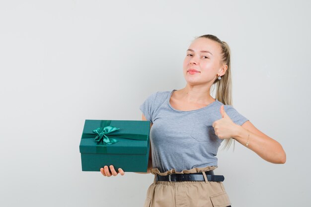 Jonge dame die huidige doos houdt, duim in t-shirt en broek toont en vreugdevol kijkt