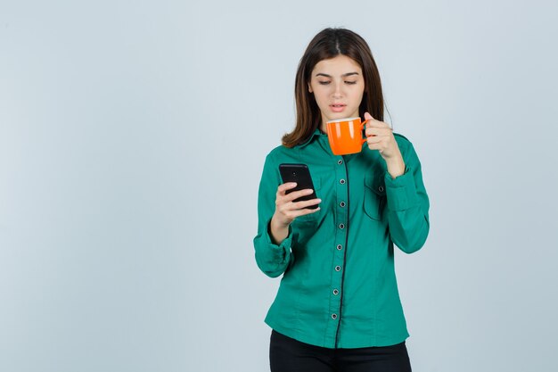 Jonge dame die in overhemd oranje kopje thee en mobiele telefoon houdt en zelfverzekerd, vooraanzicht kijkt.