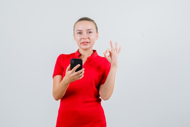 Jonge dame die in rood t-shirt mobiele telefoon met ok gebaar houdt en vrolijk kijkt