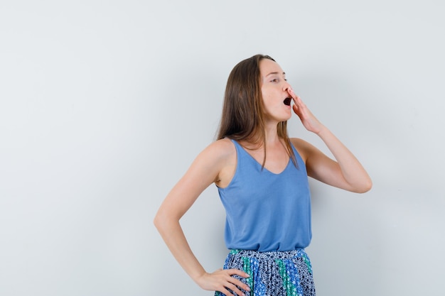 Jonge dame die met hand op mond in blauwe blouse, rok geeuwt en moe, vooraanzicht kijkt.