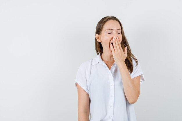 Jonge dame die met hand op mond in witte blouse geeuwt en moe kijkt.