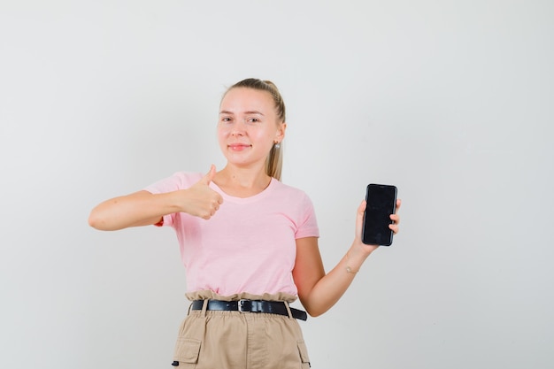 Jonge dame die mobiele telefoon houdt, duim in t-shirt en broek toont en vrolijk kijkt