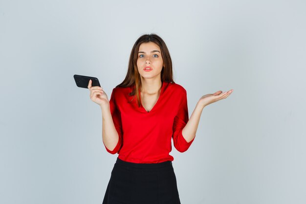 Jonge dame die mobiele telefoon houdt terwijl hulpeloos gebaar in rode blouse wordt getoond