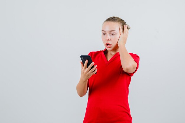 Jonge dame die mobiele telefoon in rood t-shirt bekijkt en geschokt kijkt