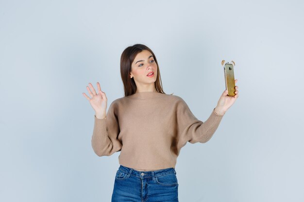 Jonge dame die mobiele telefoon vasthoudt terwijl ze palm in beige trui, jeans laat zien en er vrolijk uitziet, vooraanzicht.