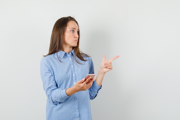 Jonge dame die mobiele telefoon vasthoudt terwijl ze weg wijst in blauw shirt en gefocust kijkt.