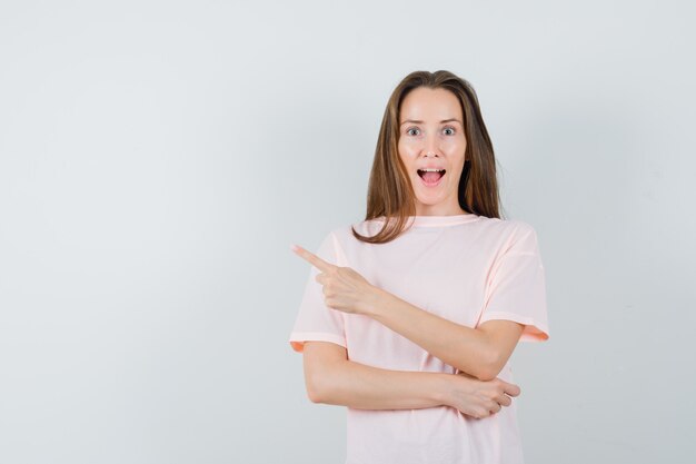 Jonge dame die naar de linkerbovenhoek in roze t-shirt wijst en verbaasd kijkt, vooraanzicht.