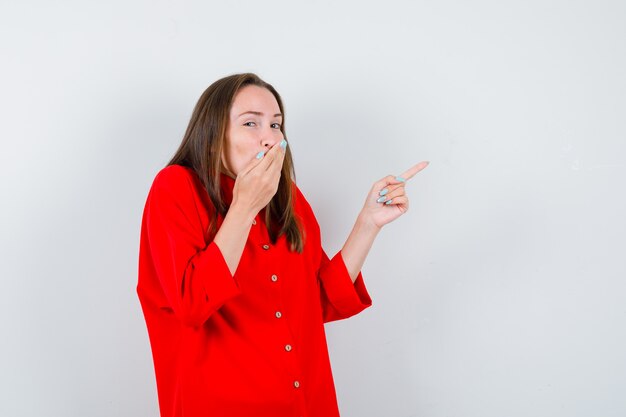 Jonge dame die naar de rechterbovenhoek wijst, de hand op de mond houdt in een rode blouse en er geamuseerd uitziet. vooraanzicht.