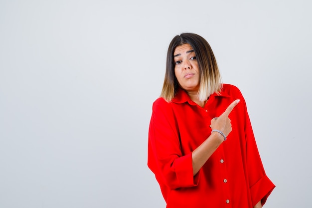 Jonge dame die naar de rechterbovenhoek wijst in een rood oversized shirt en besluiteloos kijkt, vooraanzicht.