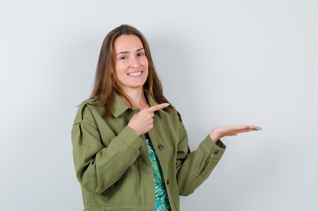 Jonge dame die naar haar handpalm wijst in een groene jas en er vrolijk uitziet, vooraanzicht.