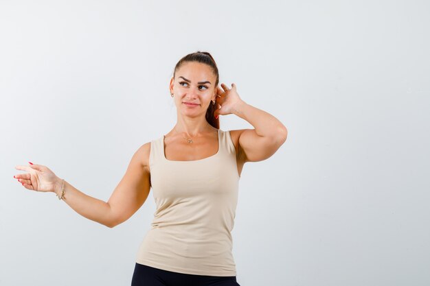 Jonge dame die naar links wijst terwijl ze haar hoofd in beige tanktop aanraakt en peinzend kijkt, vooraanzicht.