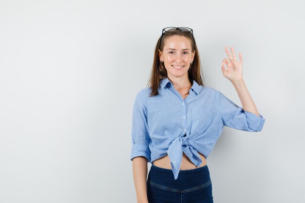 Jonge dame die ok gebaar in blauw overhemd, broek toont en tevreden kijkt.