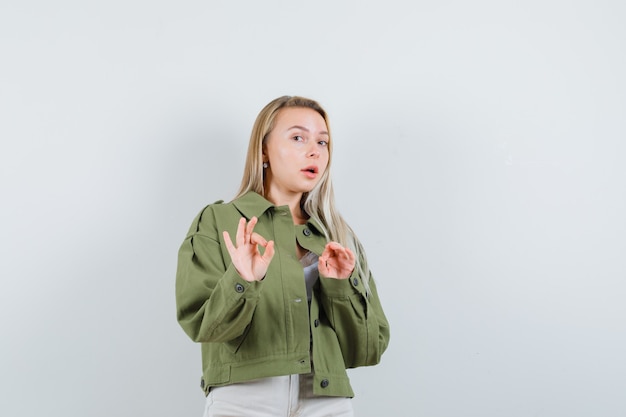 Jonge dame die ok gebaar in jasje, broek toont en zelfverzekerd, vooraanzicht kijkt.