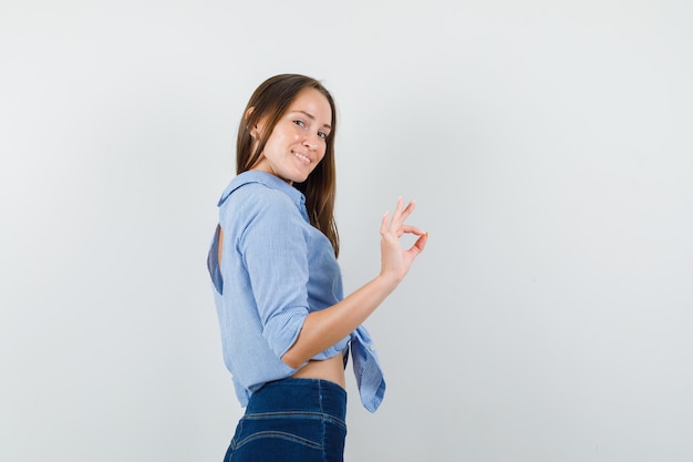 Jonge dame die ok teken in blauw overhemd, broek toont en vreugdevol kijkt.