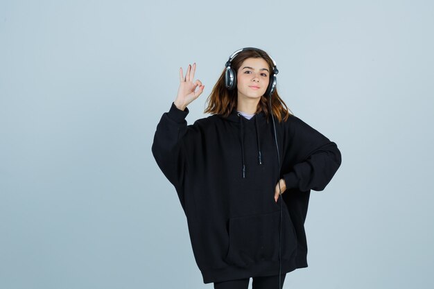 Jonge dame die ok toont terwijl ze de hand op de taille houdt in een oversized hoodie, broek en er zelfverzekerd uitziet, vooraanzicht.