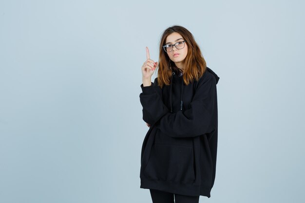 Jonge dame die omhoog wijst in oversized hoodie, broek en op zoek zelfverzekerd, vooraanzicht.