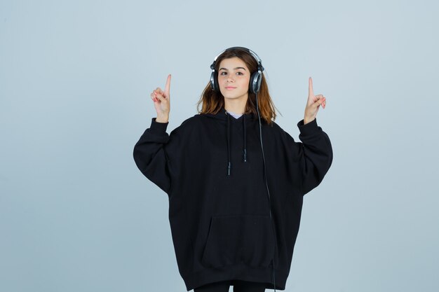 Jonge dame die omhoog wijst terwijl het luisteren naar muziek met koptelefoon in oversized hoodie, broek en op zoek zelfverzekerd, vooraanzicht.