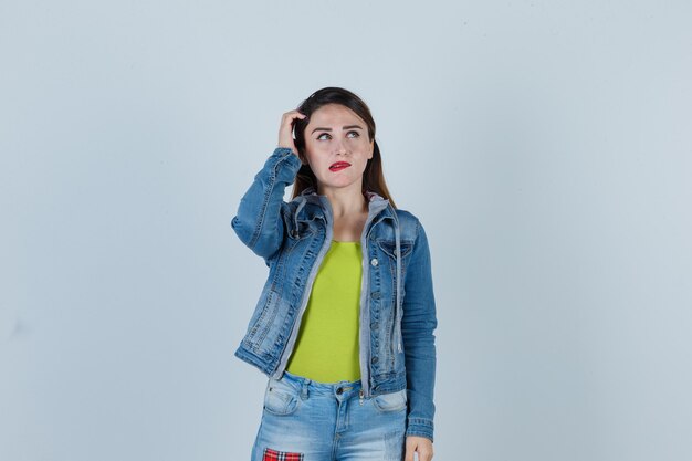 Jonge dame die opkijkt, hoofd krabt, lip bijt in denim outfit en er attent uitziet, vooraanzicht.