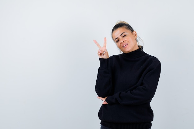 Jonge dame die overwinningsteken in zwarte sweater toont en vrolijk, vooraanzicht kijkt.