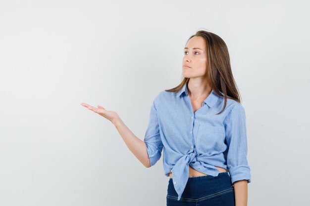 Jonge dame die palm opzij spreidt in blauw overhemd, broek en nadenkend kijkt.