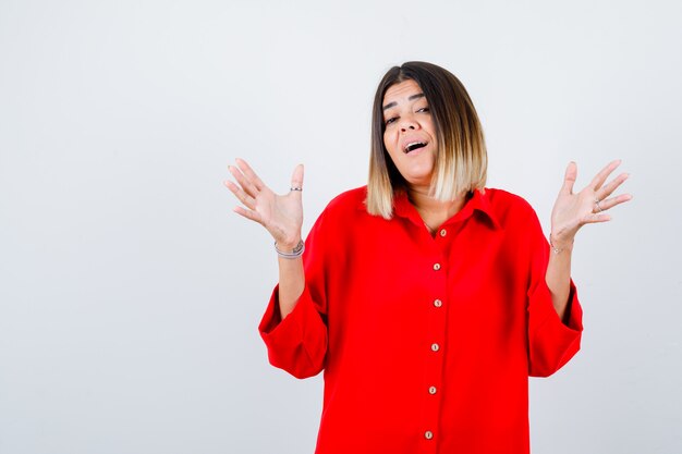 Jonge dame die palm toont terwijl ze naar de camera kijkt in een rood oversized shirt en er vrolijk uitziet, vooraanzicht.