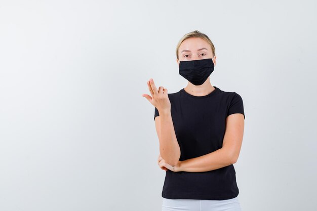 Jonge dame die pistoolgebaar toont in t-shirt, broek, medisch masker en er zelfverzekerd uitziet